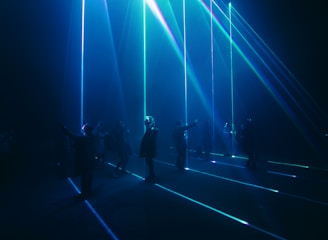 A group of people stand in a dark room with vibrant blue and green laser beams projecting vertically and horizontally around them. The atmosphere is immersive and futuristic, with shadows and light creating a dramatic effect.