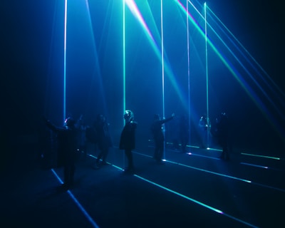 A group of people stand in a dark room with vibrant blue and green laser beams projecting vertically and horizontally around them. The atmosphere is immersive and futuristic, with shadows and light creating a dramatic effect.