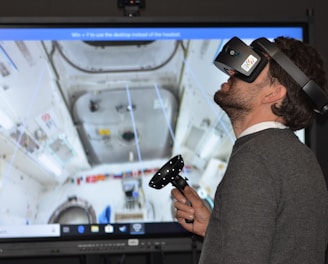 A man wearing a virtual reality headset and holding a controller is engaged in an immersive experience. The background shows a detailed screen of what appears to be the interior of a space station or similar environment, with various equipment and panels visible.