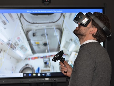 A man wearing a virtual reality headset and holding a controller is engaged in an immersive experience. The background shows a detailed screen of what appears to be the interior of a space station or similar environment, with various equipment and panels visible.