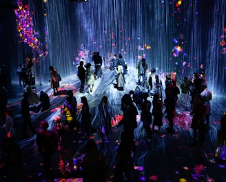 A crowd of people stands in a dark room illuminated by a vibrant digital projection. The projection features cascading lines resembling a waterfall, interspersed with colorful patches resembling flowers or fireworks. The light reflects off the floor, creating an immersive and interactive environment.