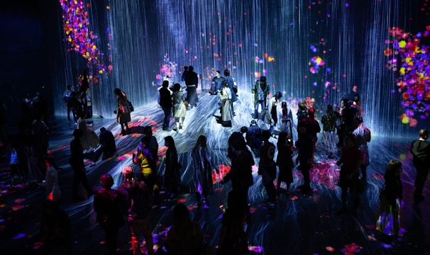 A crowd of people stands in a dark room illuminated by a vibrant digital projection. The projection features cascading lines resembling a waterfall, interspersed with colorful patches resembling flowers or fireworks. The light reflects off the floor, creating an immersive and interactive environment.