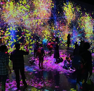 A vibrant and immersive digital art installation with bright, colorful light projections resembling flowers and abstract patterns covering the walls and floor. People are interacting with the environment, some sitting and others standing or walking around, creating an interactive and dynamic atmosphere.