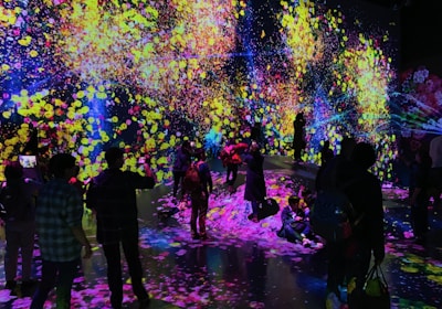 A vibrant and immersive digital art installation with bright, colorful light projections resembling flowers and abstract patterns covering the walls and floor. People are interacting with the environment, some sitting and others standing or walking around, creating an interactive and dynamic atmosphere.