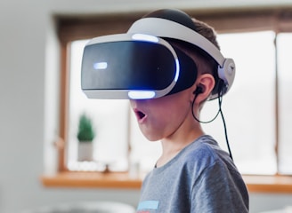 A young boy wearing a virtual reality headset appears to be engaged and surprised. The environment is indoors, with soft lighting and a blurred background. The boy is wearing a casual grey T-shirt, and the headset has glowing blue lights.