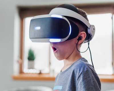 A young boy wearing a virtual reality headset appears to be engaged and surprised. The environment is indoors, with soft lighting and a blurred background. The boy is wearing a casual grey T-shirt, and the headset has glowing blue lights.