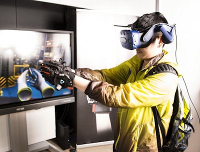 A person is engaged in a virtual reality experience, wearing a VR headset and gloves. They are facing a large screen displaying a simulated environment with tubes and industrial elements. The individual is wearing a yellow jacket and a backpack, appearing focused on the VR interaction.