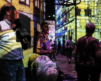 People are standing and walking in a dimly lit room with colorful digital projections on the walls, creating an immersive art experience. The projections depict a detailed building facade. The atmosphere appears lively with several individuals engaging with the artwork.