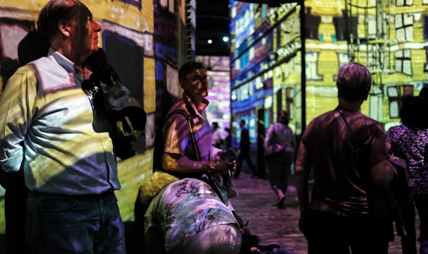People are standing and walking in a dimly lit room with colorful digital projections on the walls, creating an immersive art experience. The projections depict a detailed building facade. The atmosphere appears lively with several individuals engaging with the artwork.