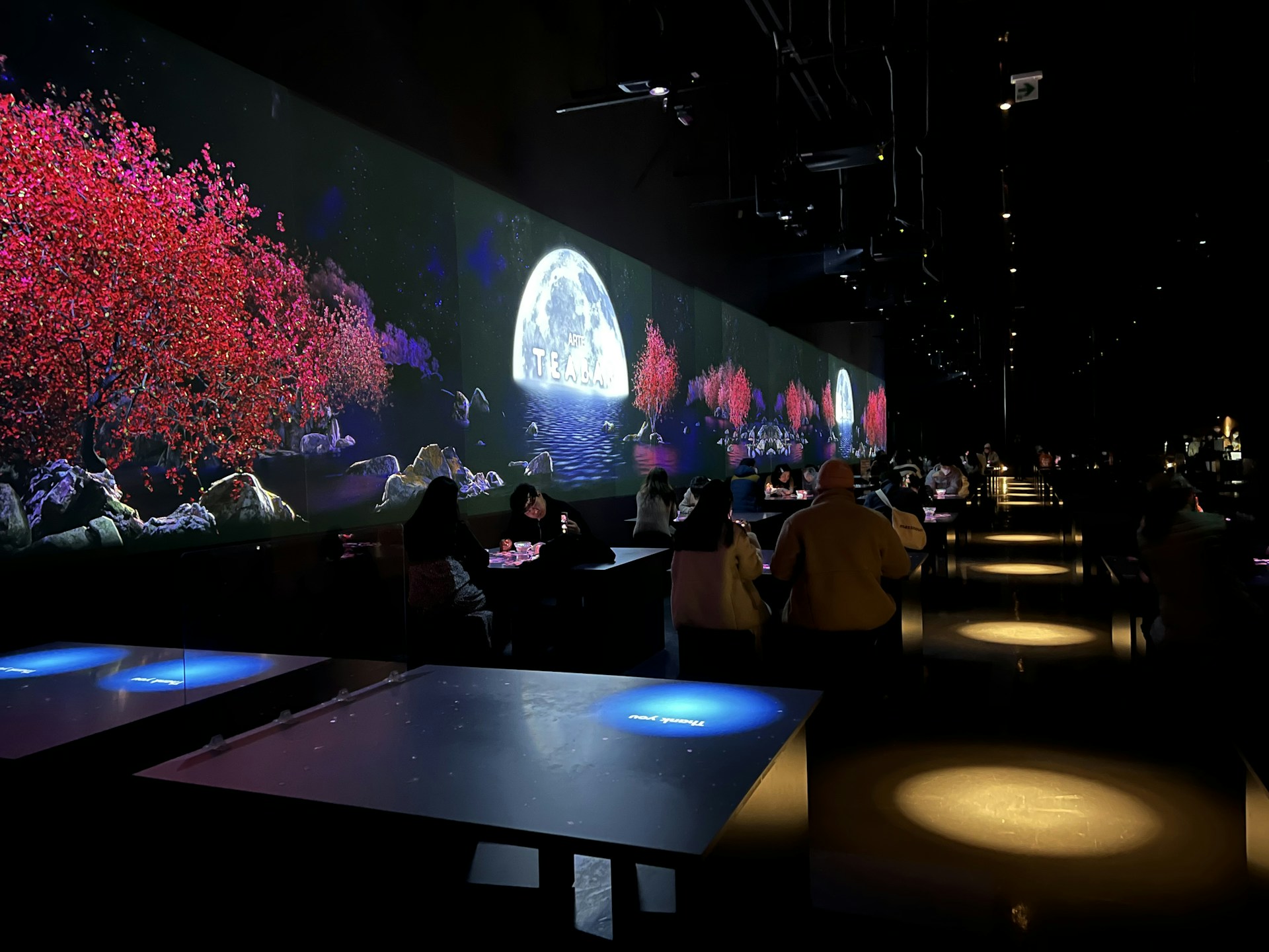 The image features a dimly lit indoor setting with long tables and people seated. A vibrant digital display on the wall shows a serene scene with red-leaved trees, rocky terrain, and a full moon over water, suggesting an immersive experience.