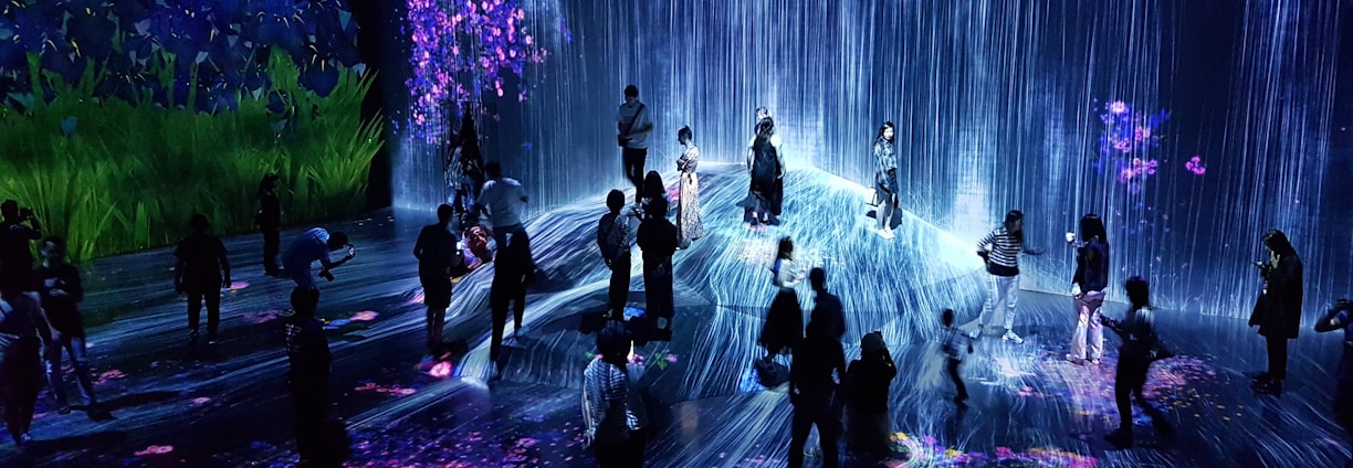 A group of people are standing and walking inside an immersive art installation with flowing digital streams and vibrant floral projections on the walls and floor. The environment appears interactive, with the participants seemingly engaged with the digital elements around them.