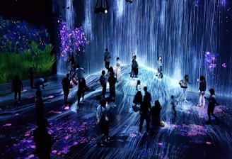 A group of people are standing and walking inside an immersive art installation with flowing digital streams and vibrant floral projections on the walls and floor. The environment appears interactive, with the participants seemingly engaged with the digital elements around them.
