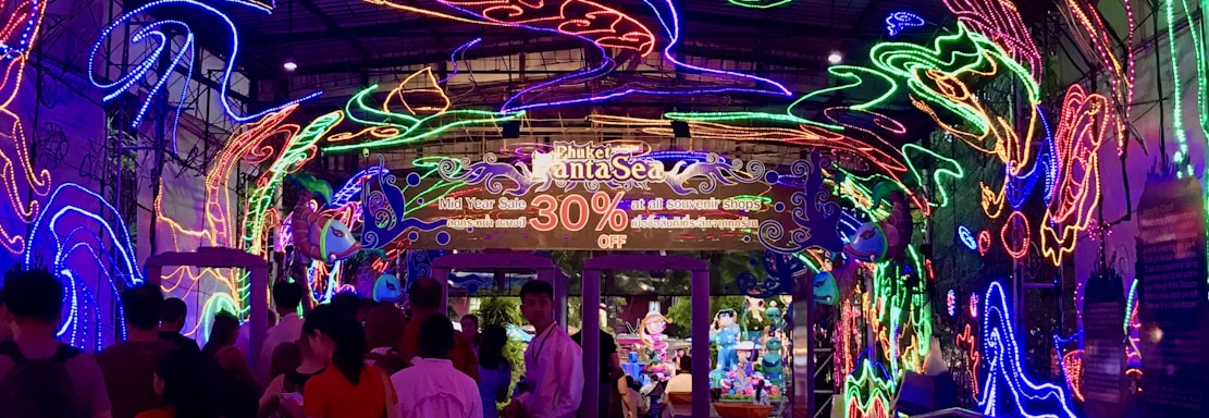 A vibrant and colorful space filled with neon light designs on the ceiling and walls, creating an immersive atmosphere. People are gathered in a queue, likely waiting to enter an attraction. The environment is reminiscent of an aquarium or theme park setting, with signs and decorations enhancing the lively mood.