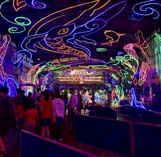 A vibrant and colorful space filled with neon light designs on the ceiling and walls, creating an immersive atmosphere. People are gathered in a queue, likely waiting to enter an attraction. The environment is reminiscent of an aquarium or theme park setting, with signs and decorations enhancing the lively mood.
