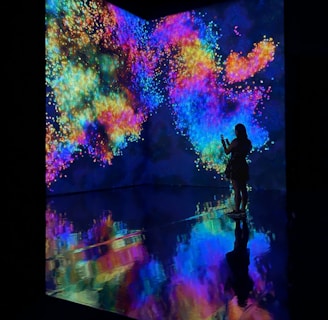 An immersive digital art installation features vibrant, swirling colors projected onto walls. A person stands in silhouette against this vivid backdrop, taking a photo or video. The floor reflects the colorful patterns, creating a mirrored effect.