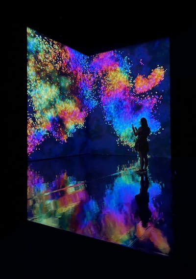 An immersive digital art installation features vibrant, swirling colors projected onto walls. A person stands in silhouette against this vivid backdrop, taking a photo or video. The floor reflects the colorful patterns, creating a mirrored effect.