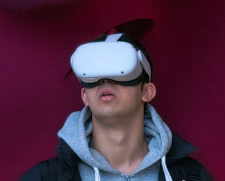 A person wearing a virtual reality headset appears to be engaged or immersed in the experience. They are dressed in a casual outfit consisting of a hoodie and a jacket, with a light-colored background offering a contrast to the darker tones of the clothing.
