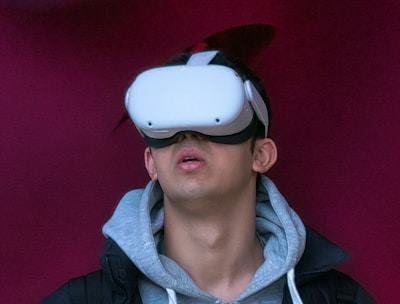 A person wearing a virtual reality headset appears to be engaged or immersed in the experience. They are dressed in a casual outfit consisting of a hoodie and a jacket, with a light-colored background offering a contrast to the darker tones of the clothing.