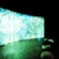 A large, curved screen displays vibrant, abstract patterns with dominant shades of blue and green, creating a surreal and immersive atmosphere. The room is dimly lit, enhancing the luminous effect of the screen. In the foreground, there are several round cushions placed on a concrete floor.