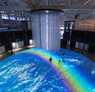 An indoor scene with a large circular projection on the floor depicting an ocean with a vibrant rainbow. Three people are standing on the projection, which creates an immersive effect. A cylindrical installation with lights hangs from the ceiling, adding to the atmosphere.