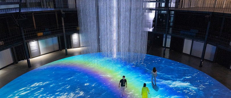 An indoor scene with a large circular projection on the floor depicting an ocean with a vibrant rainbow. Three people are standing on the projection, which creates an immersive effect. A cylindrical installation with lights hangs from the ceiling, adding to the atmosphere.